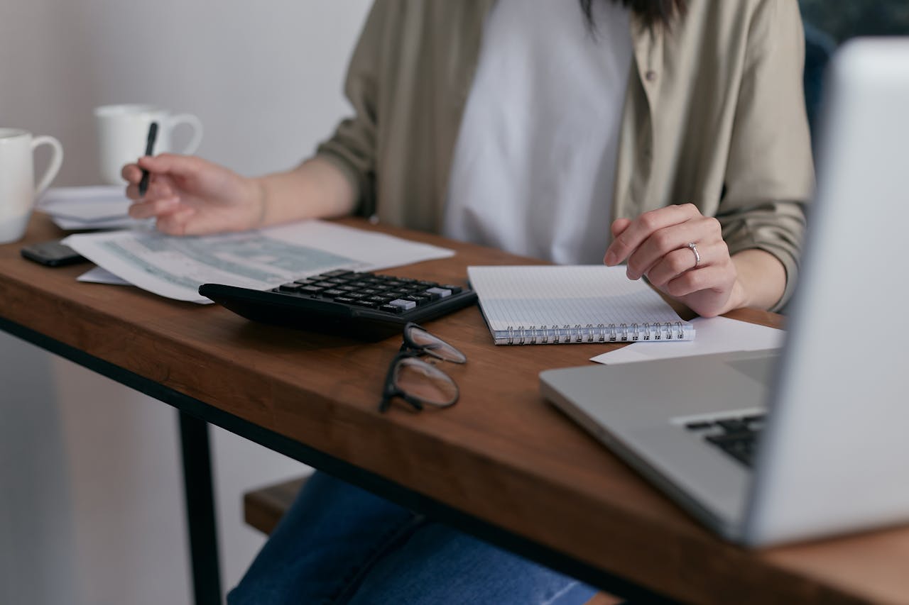 gallery-02 A woman manages finances at home, using a laptop and calculator on a wooden desk.