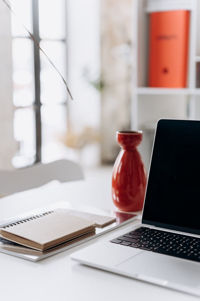 trusted-partner A minimalist home office setup featuring a laptop, notebook, and a red vase on a white desk.
