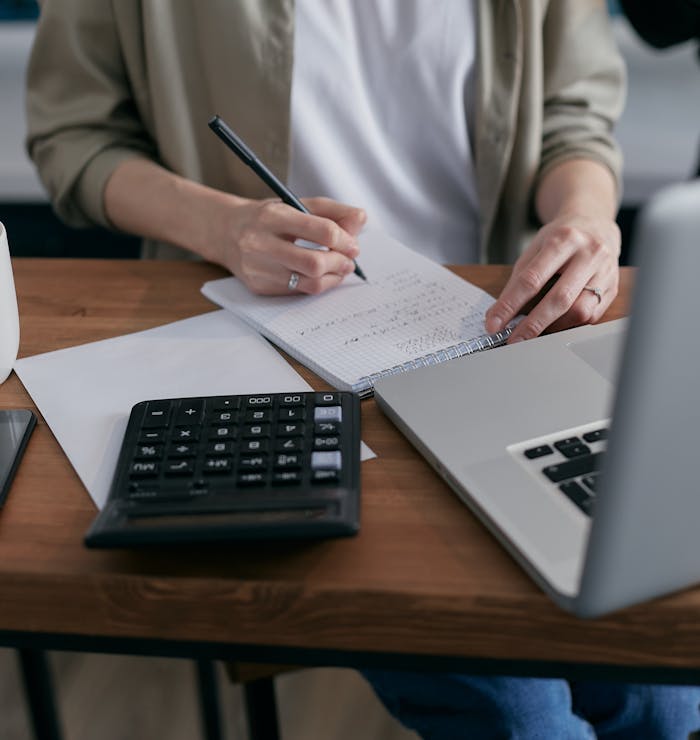 gallery-04 A woman writes financial calculations in a notebook, using a calculator and laptop at a wooden desk.